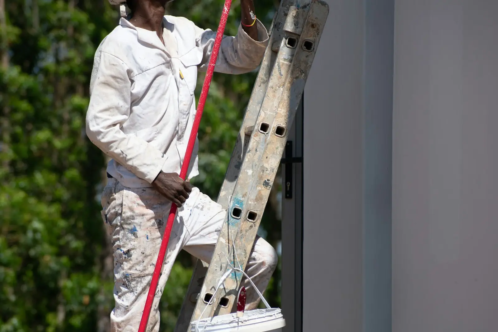 Painter and decorator in paint-spattered overalls climbing a ladder with a roller pole